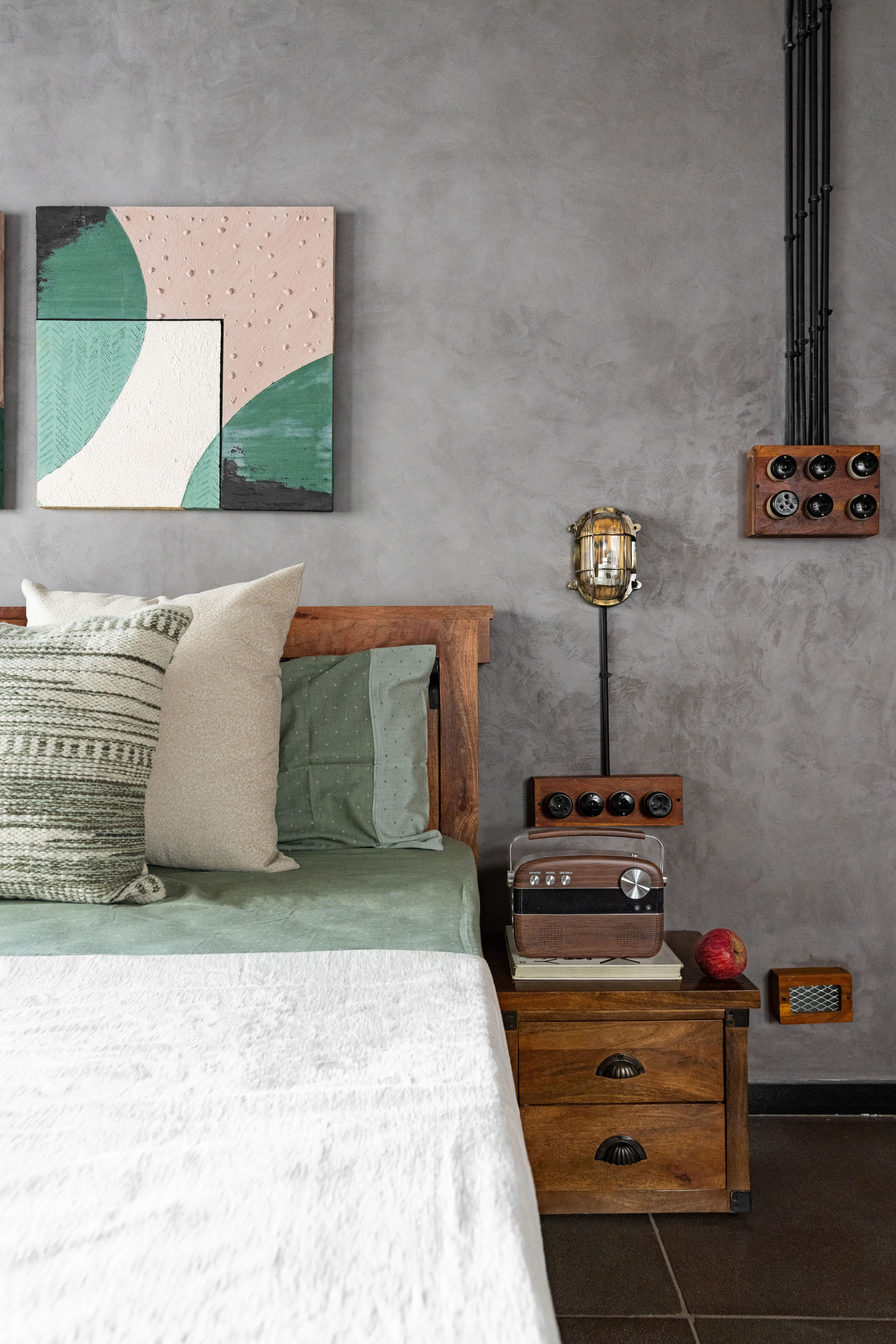 Bedside in the second bedroom: sage-green bedding and textured cushions on a timber bed, abstract relief panels in green, blush and cream above, a caged lamp on a stand, and a retro radio on a stack of books beside a red apple on the nightstand.
